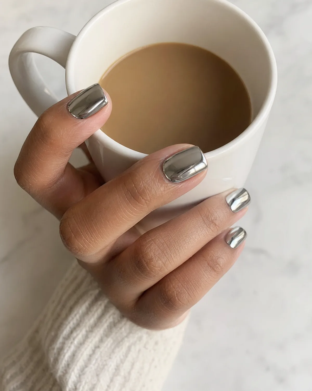 Three-quarter overhead view of a hand with short square chrome silver acrylic nails wrapped around a white coffee mug