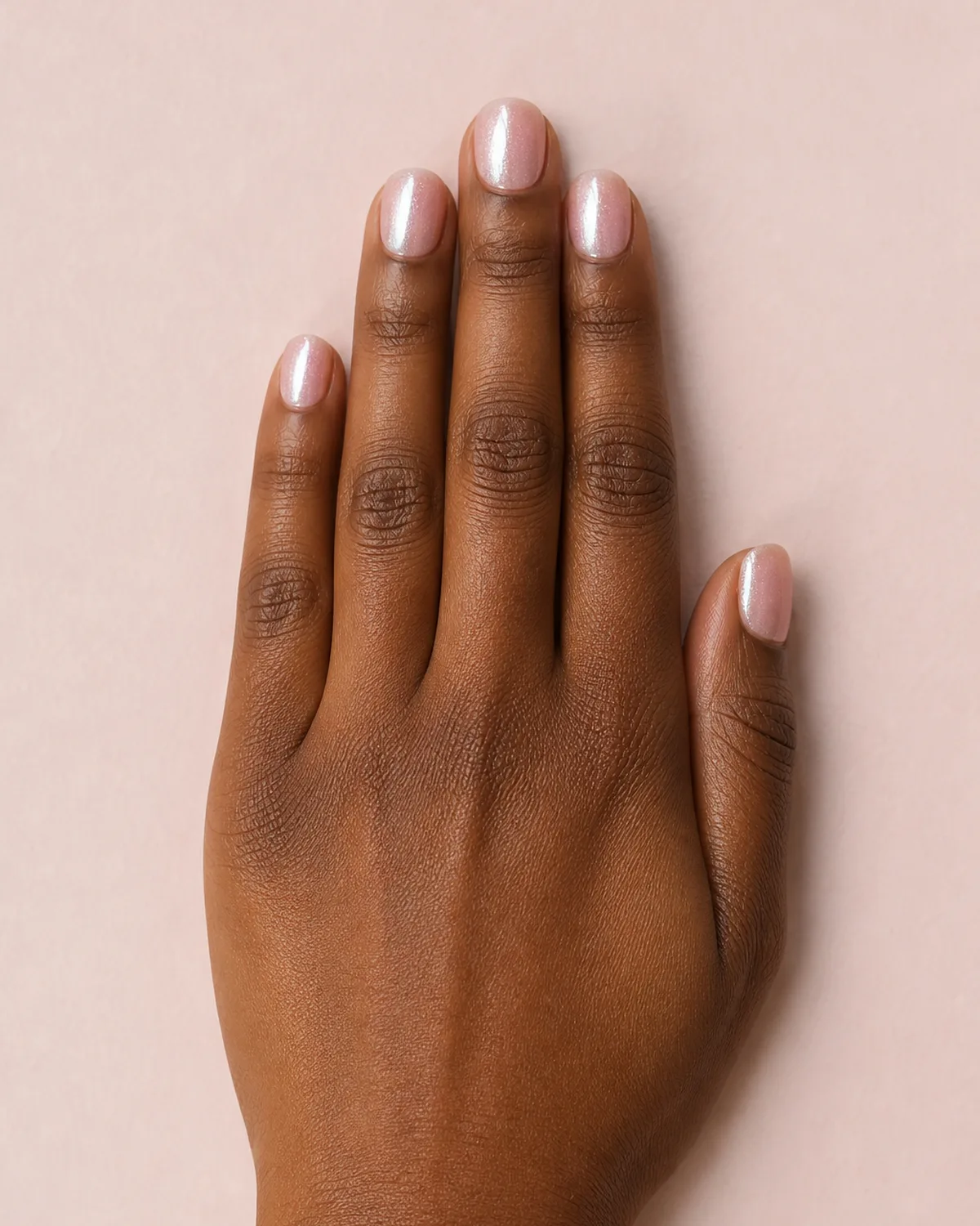 Overhead view of a hand with short round acrylic nails in glazed donut pearlescent shimmer on a pale pink surface