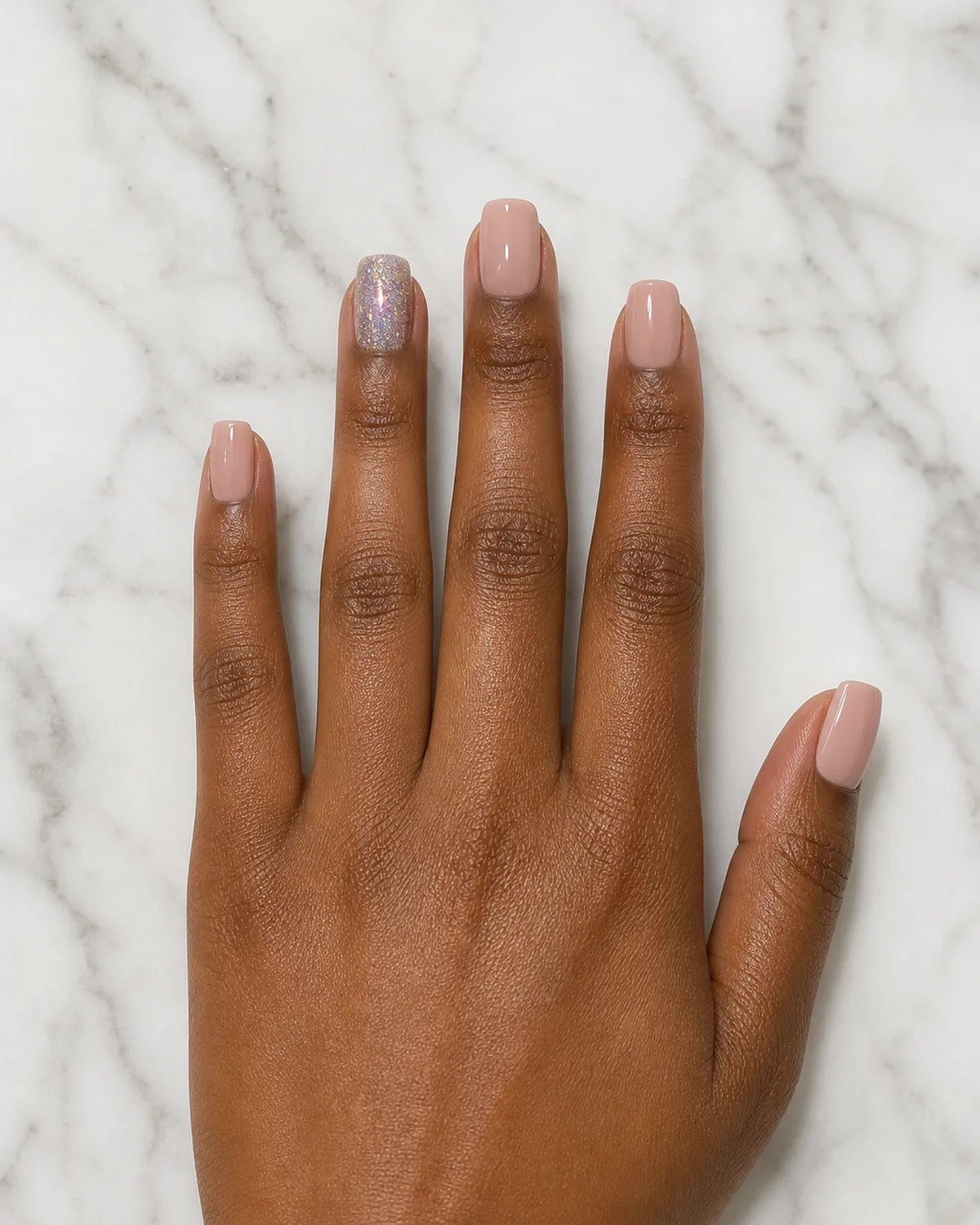 Overhead view of a hand on marble with short coffin acrylic nails, four nude and one holographic glitter ring finger
