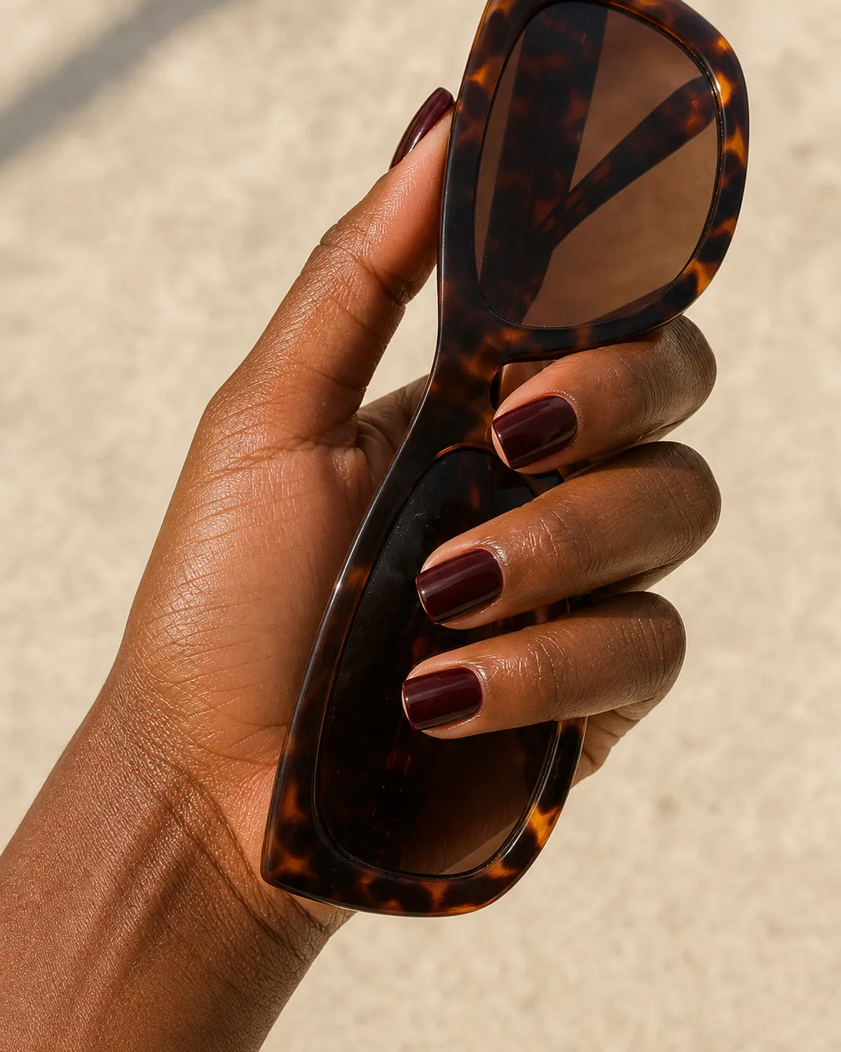Three-quarter view of a hand with short square acrylic nails in deep burgundy holding tortoiseshell sunglasses