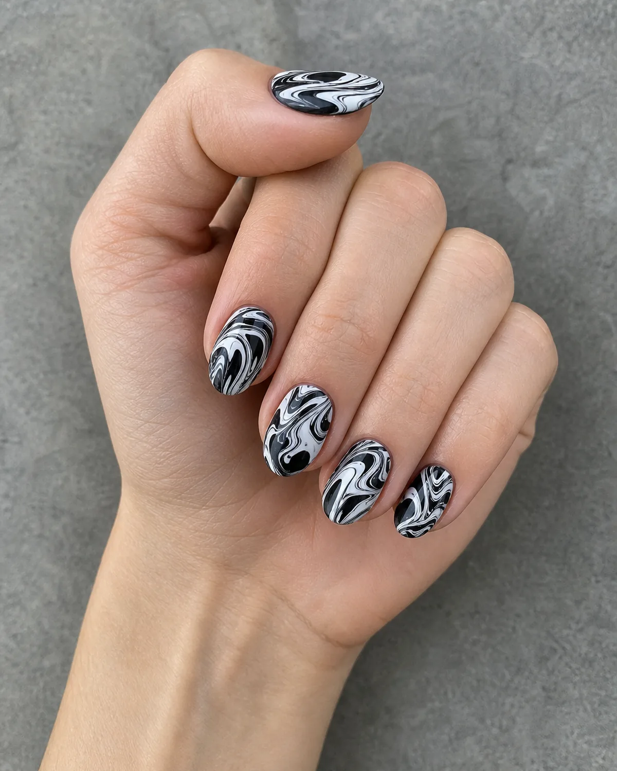 Close-up of short almond nails with a black and white high-contrast water marble swirl, on light skin resting on grey stone