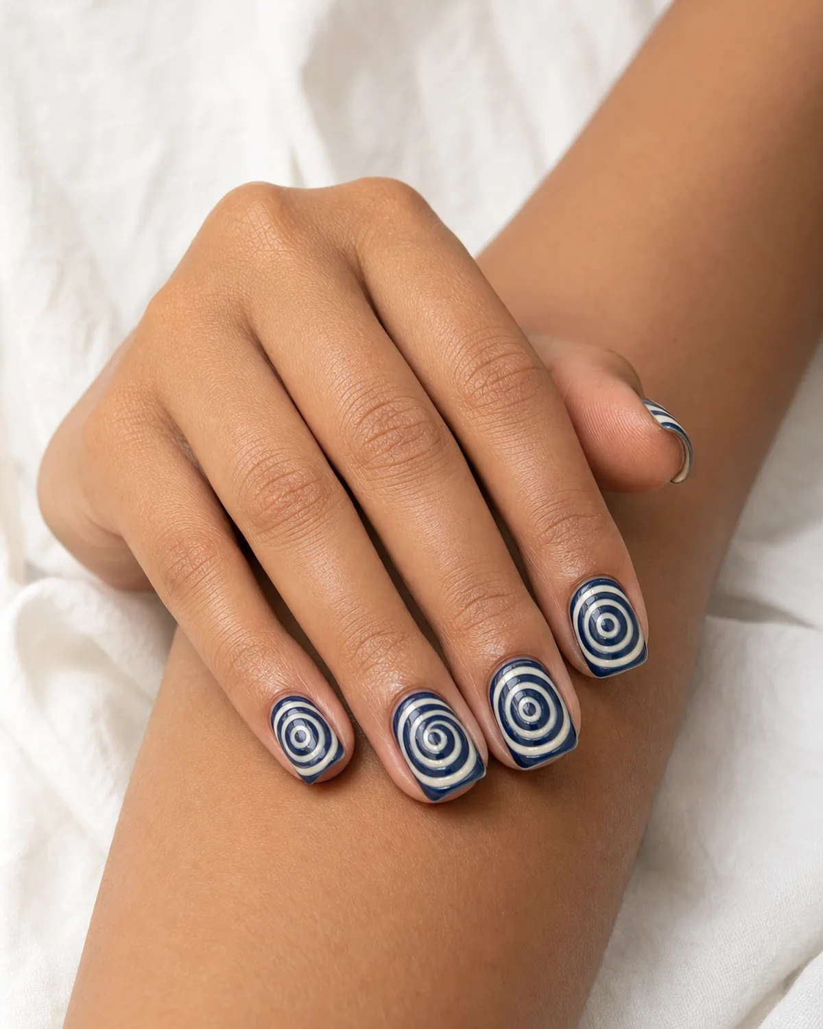 Close-up of medium square nails with a tight concentric-ring water marble pattern in navy and cream, on medium skin resting on white linen