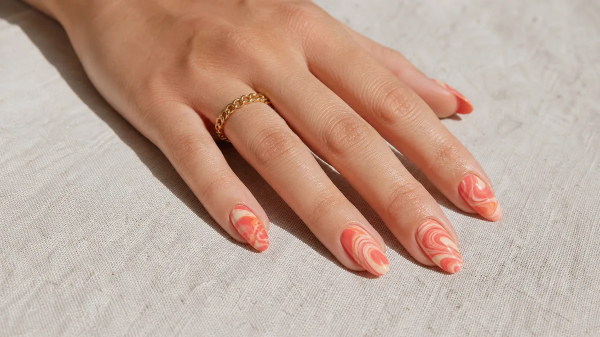 A hero close-up of medium almond nails in a warm two-tone coral-and-cream water marble swirl, on light-medium skin resting on pale linen with soft natural daylight
