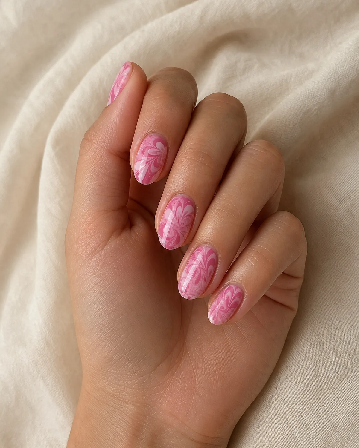Close-up of medium oval nails with a feathered light-pink and rose floral bloom water marble pattern, on light-medium skin resting on cream fabric