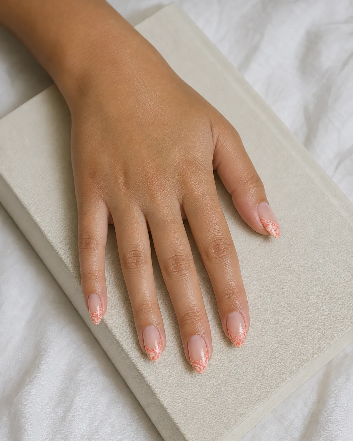 Close-up of medium almond nails with a sheer nude base and a coral-cream water marble swirl confined to the French tip area, on medium skin resting on white linen