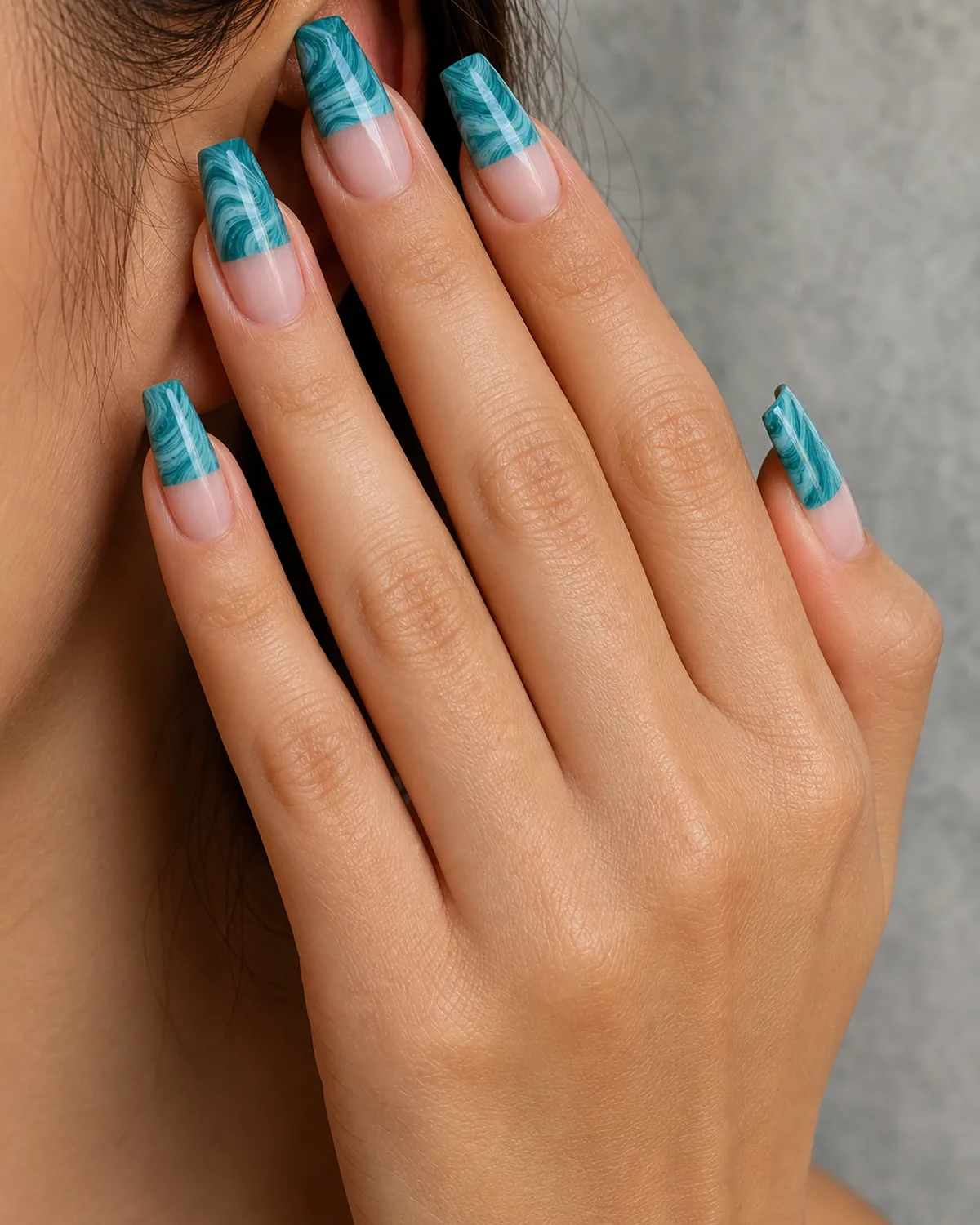 Close-up of medium coffin nails with a half-dip teal water marble pattern leaving the cuticle area bare, on light-medium skin resting on grey stone