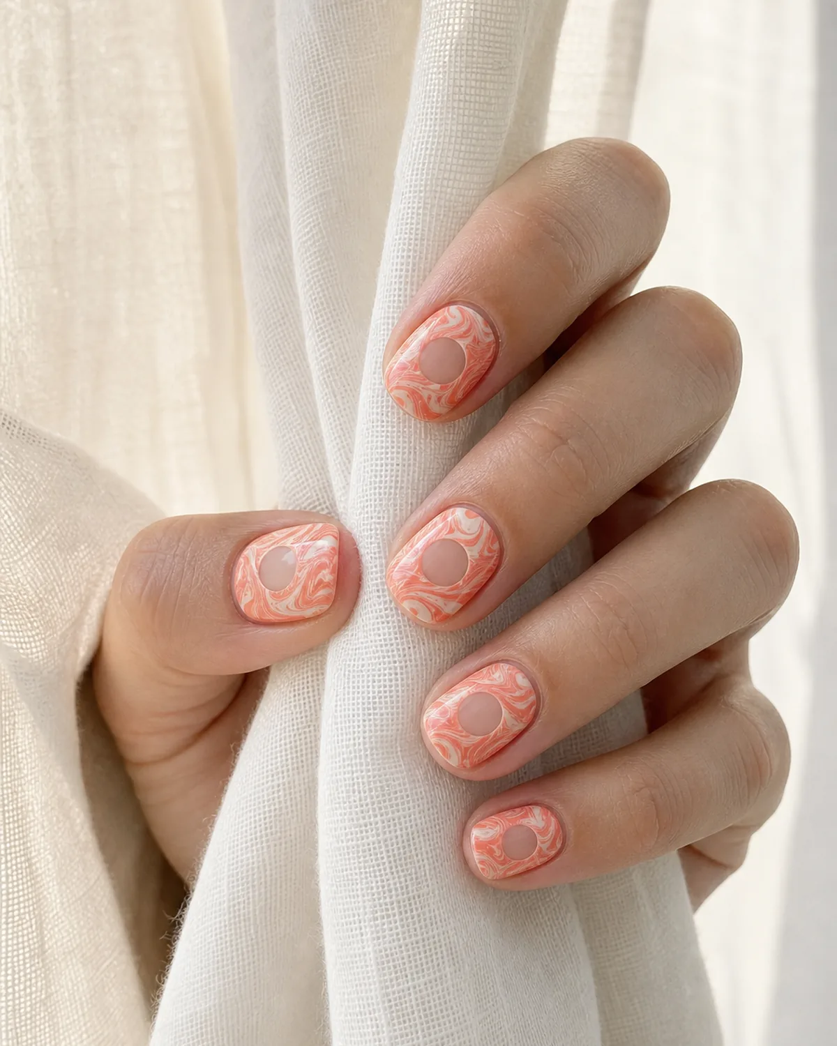 Close-up of medium round nails with a negative-space water marble swirl in coral and cream framing a bare center circle, on light-medium skin resting on pale linen