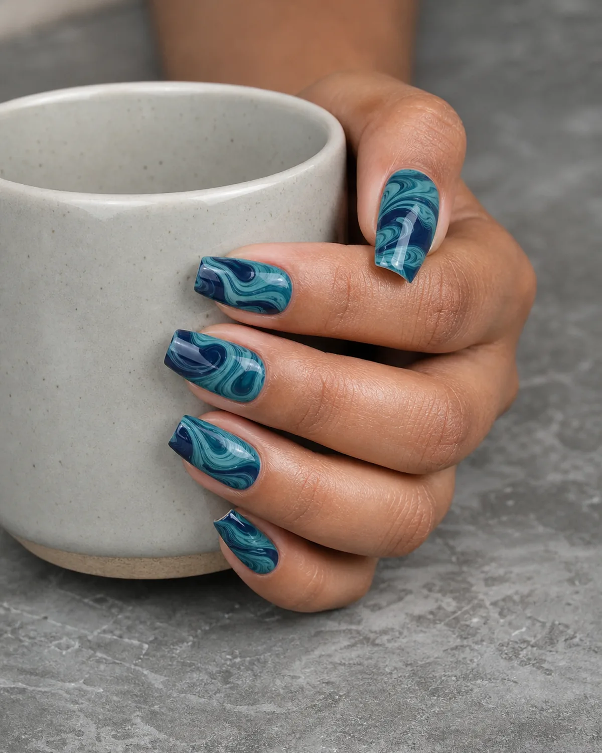 Close-up of medium coffin nails with a teal and deep navy water marble swirl, on medium skin resting on grey stone