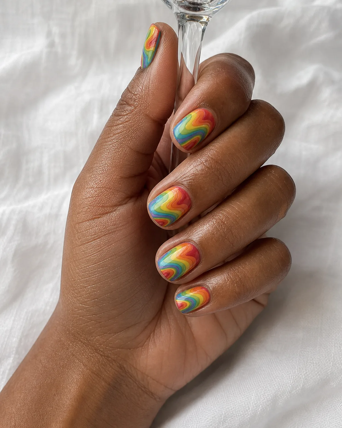 Close-up of short square nails with a rainbow five-color water marble swirl, on medium-dark skin resting on white linen