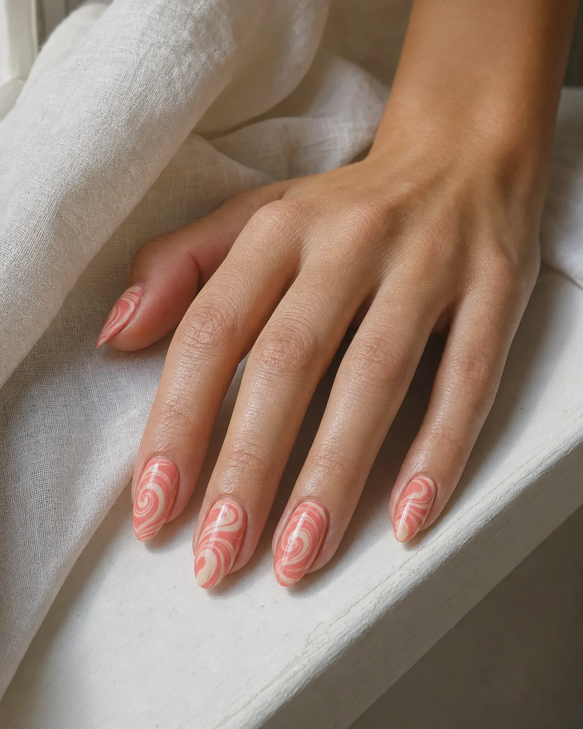 Close-up of medium almond nails with a classic two-tone coral and cream water marble swirl, on light-medium skin resting on pale linen