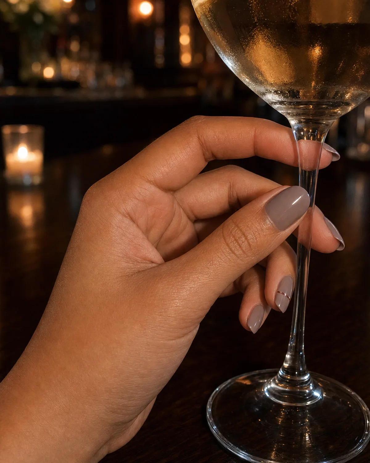 Soft taupe nails with one thin rose-gold accent line on the ring finger, medium oval, hand near a wine glass stem