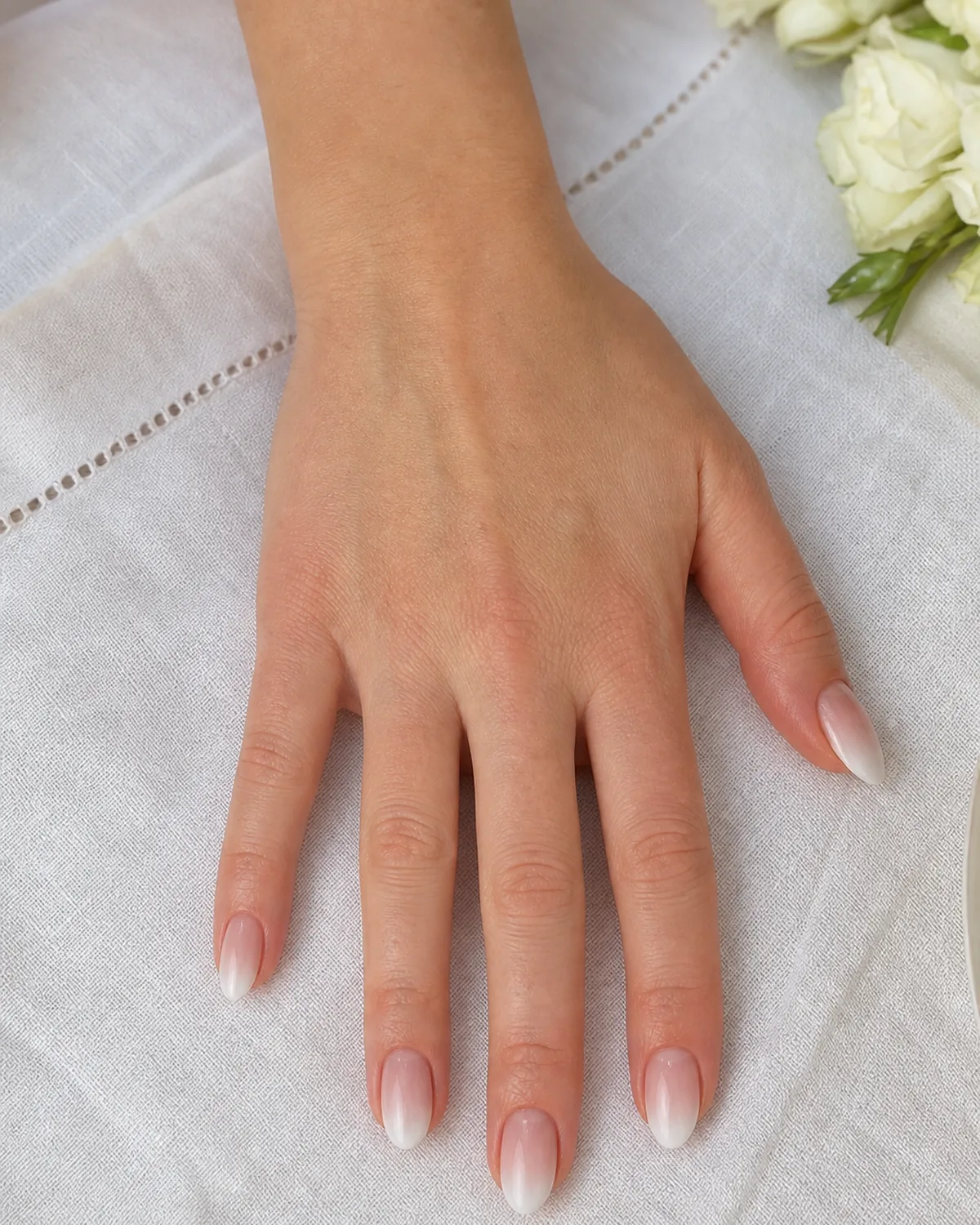 Close-up of medium almond nails with a baby-boomer gradient from soft white tip to sheer warm pink base, on fair skin on a white linen napkin