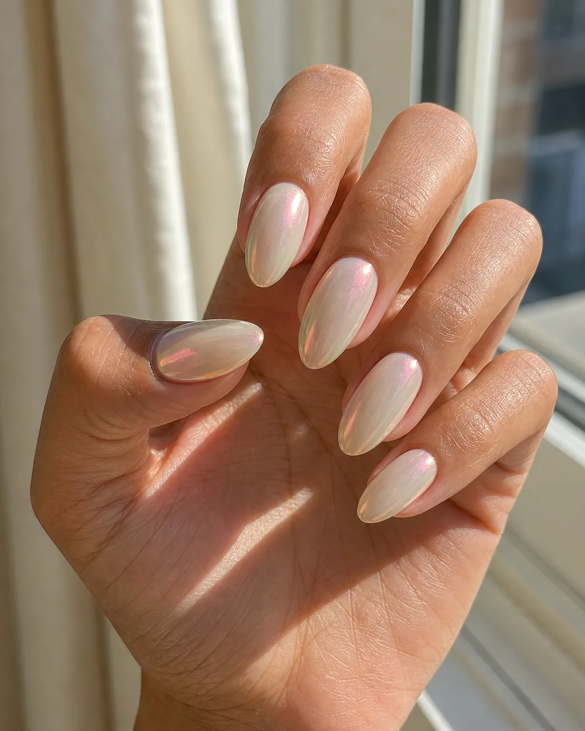 Close-up of medium almond nails in an iridescent glazed finish on an ivory base, shifting pink-to-gold, near a sunlit window