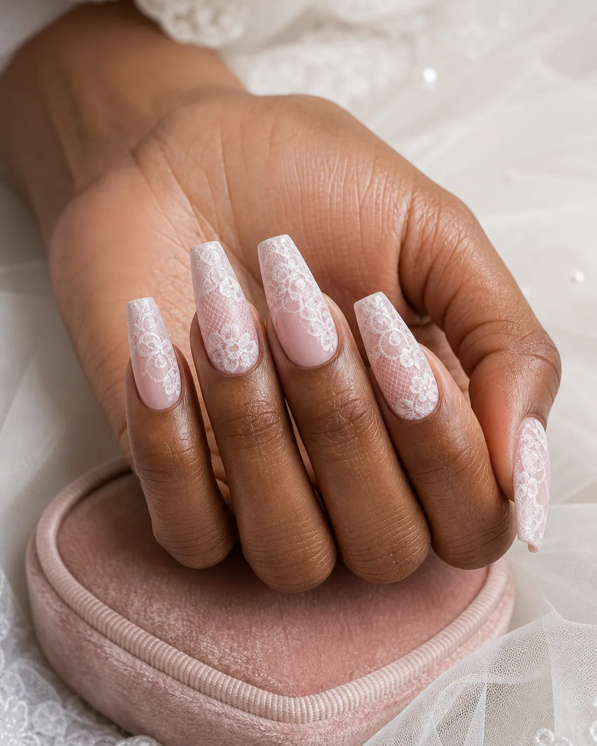 Close-up of long coffin nails with delicate white lace nail art over a sheer pink base, on medium-dark skin near a blush velvet ring box