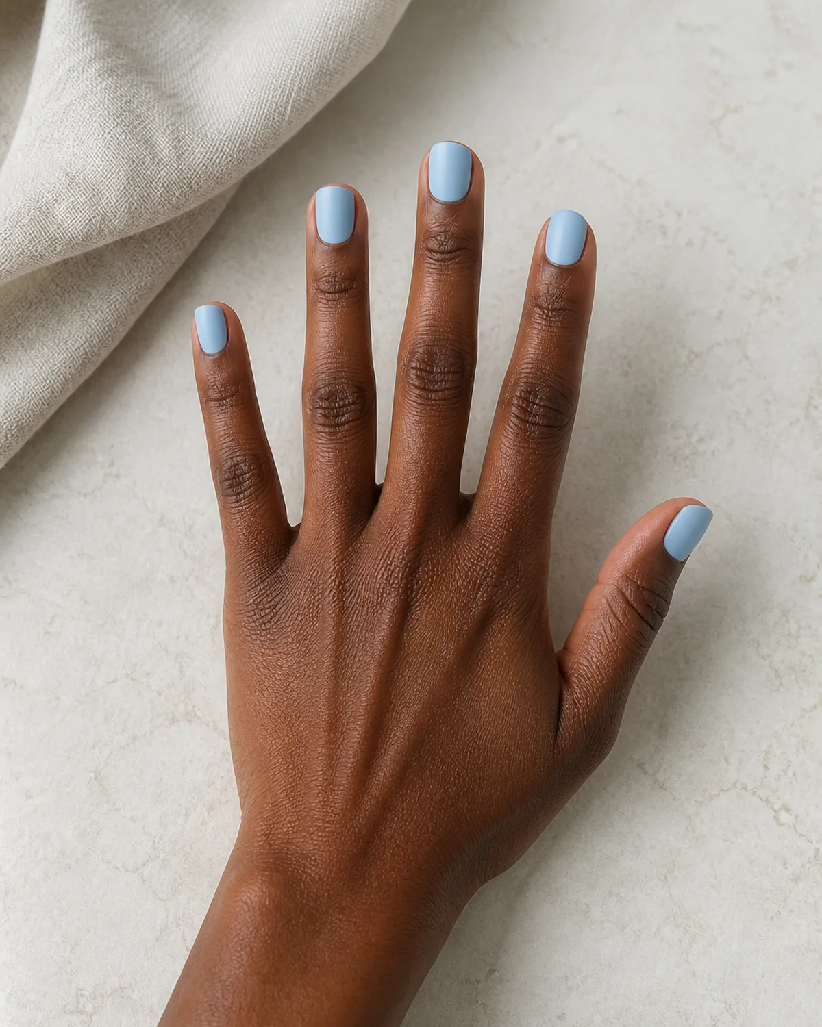 Single hand with powder blue matte nails on short square shape with fingers spread in a fan on a pale surface beside cream linen