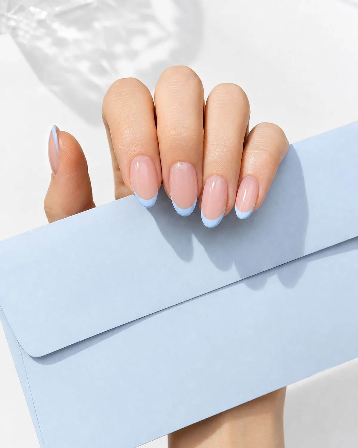 Single hand with baby blue french tip nails on medium almond shape holding a pale blue envelope against a white surface
