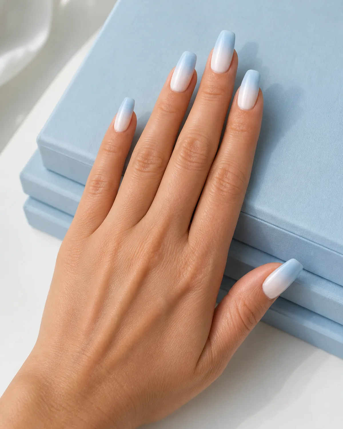 Single hand with baby blue ombre nails on coffin shape transitioning from white at cuticle to sky blue at tip resting beside pastel blue hardcover books