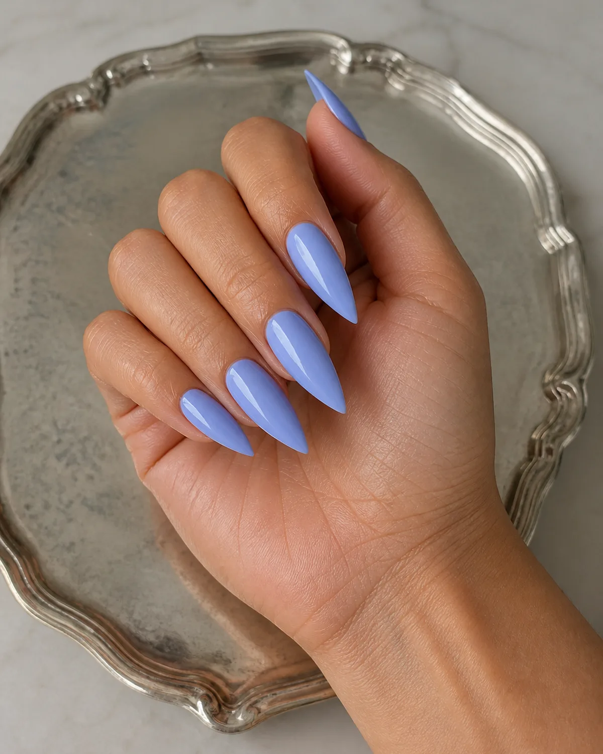Single hand with periwinkle-blue nails on stiletto shape with fingers resting on a silver jewelry tray