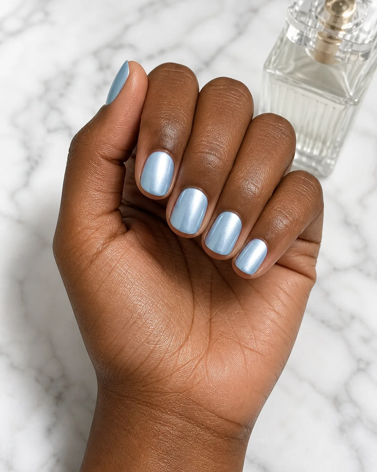 Single hand with baby blue chrome nails with pearlescent shimmer on short round shape in a loose fist on white marble beside a glass perfume bottle