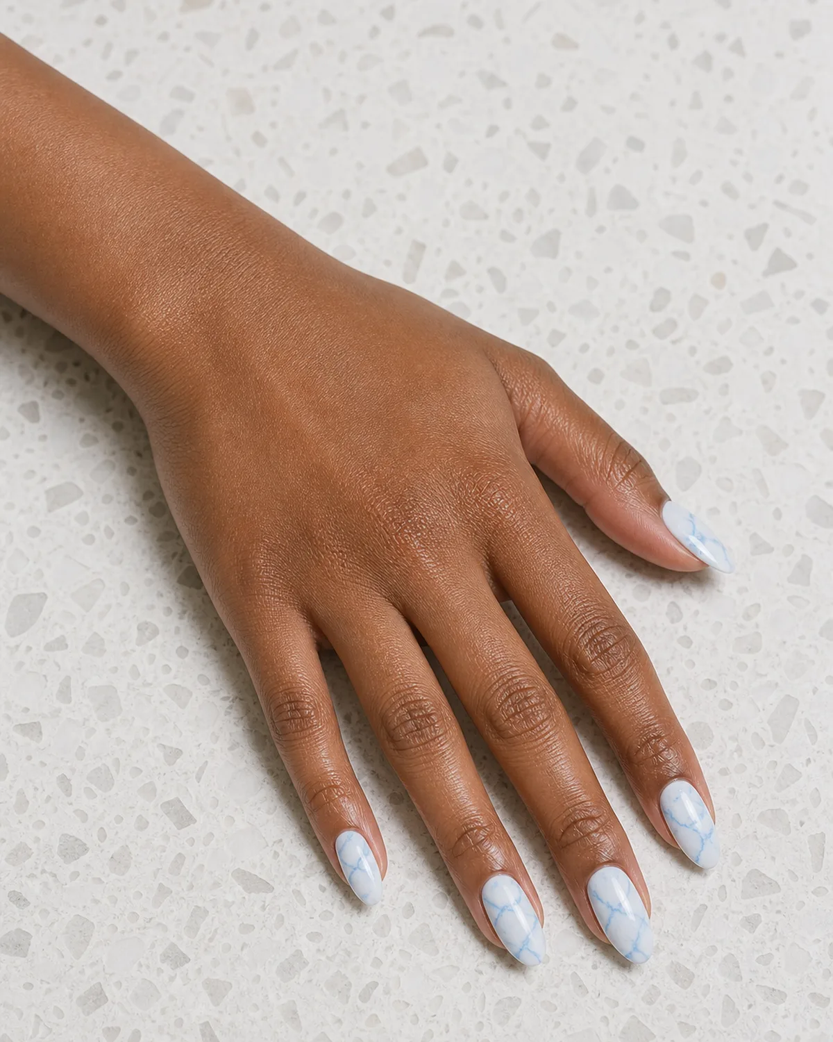 Single hand with pale blue marble nails on almond shape with white base and soft blue veining resting on a white terrazzo surface