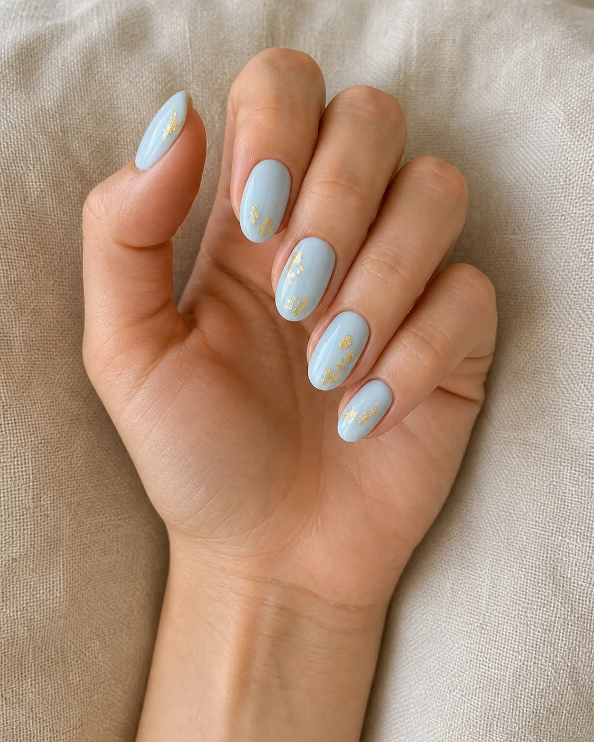 Single hand with baby blue nails with scattered gold leaf foil accents on medium oval shape resting on a pale beige linen cushion