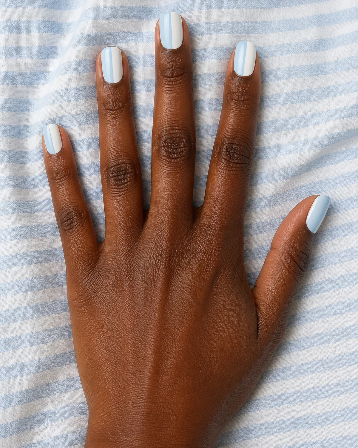 Single hand with baby blue and white color block nails in geometric half-and-half split on medium square shape splayed flat on pale blue-and-white striped fabric