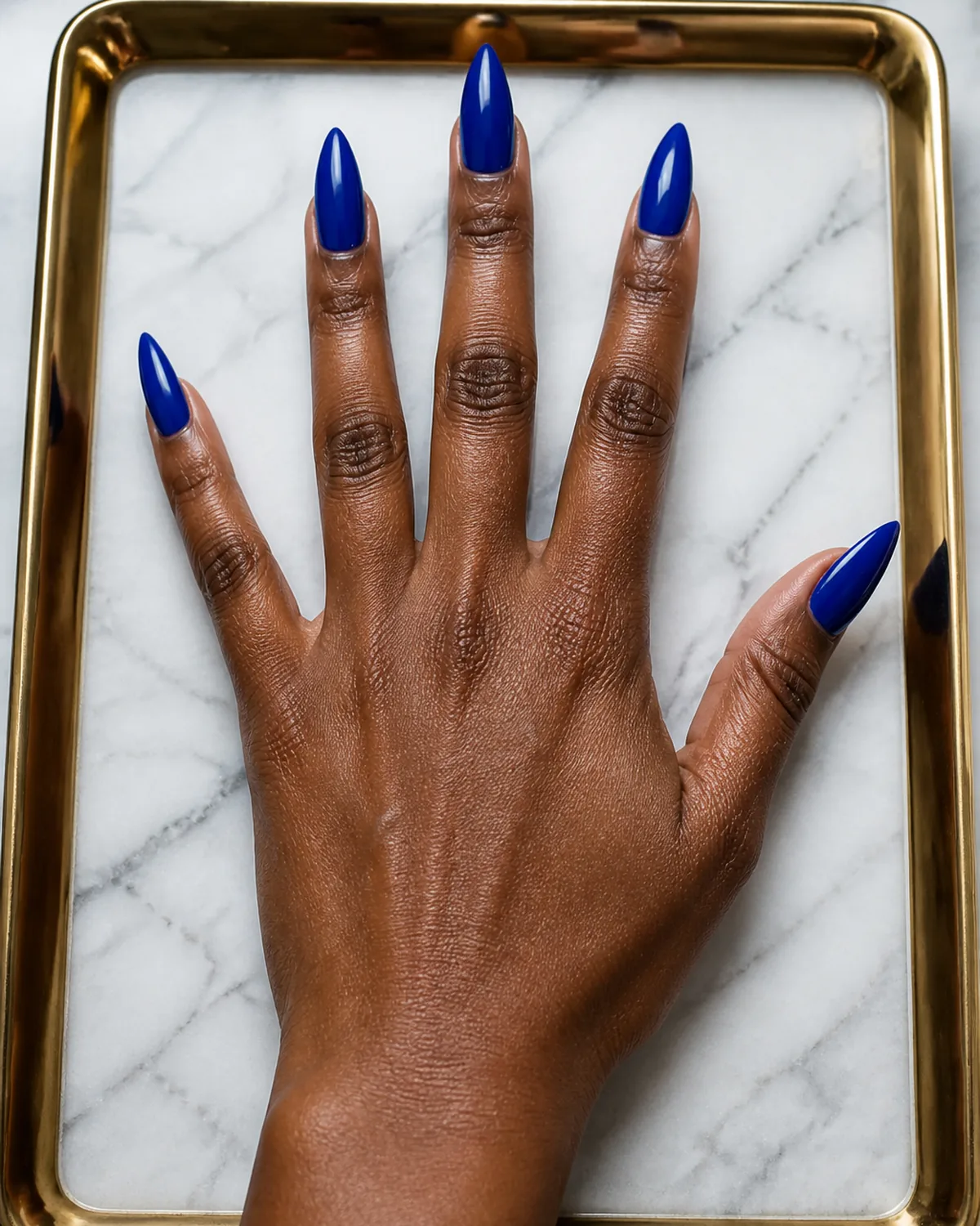 Single hand with saturated royal blue nails on stiletto shape with fingers fanned on a gold-framed tray on white marble