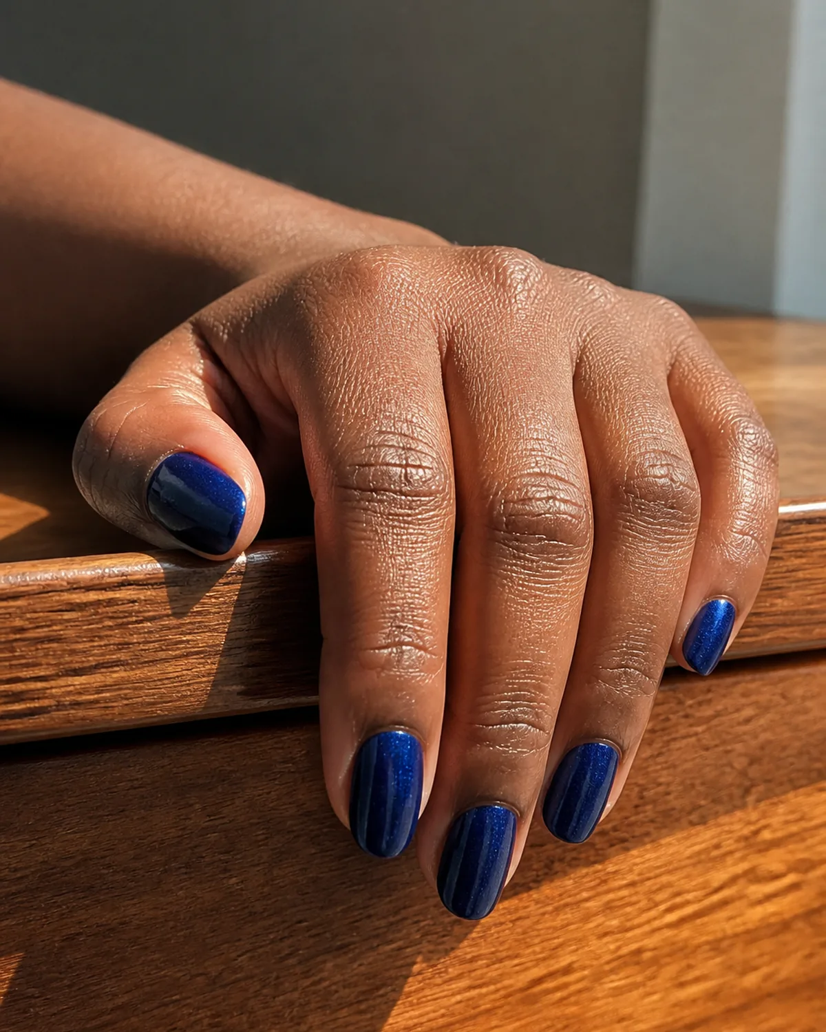 Single hand with deep sapphire blue shimmer nails on oval shape curled over the edge of a wooden vanity ledge