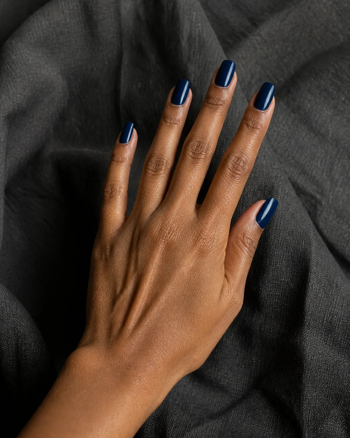 Single hand with deep inky indigo blue nails on coffin shape reaching diagonally with fingers relaxed against dark gray linen