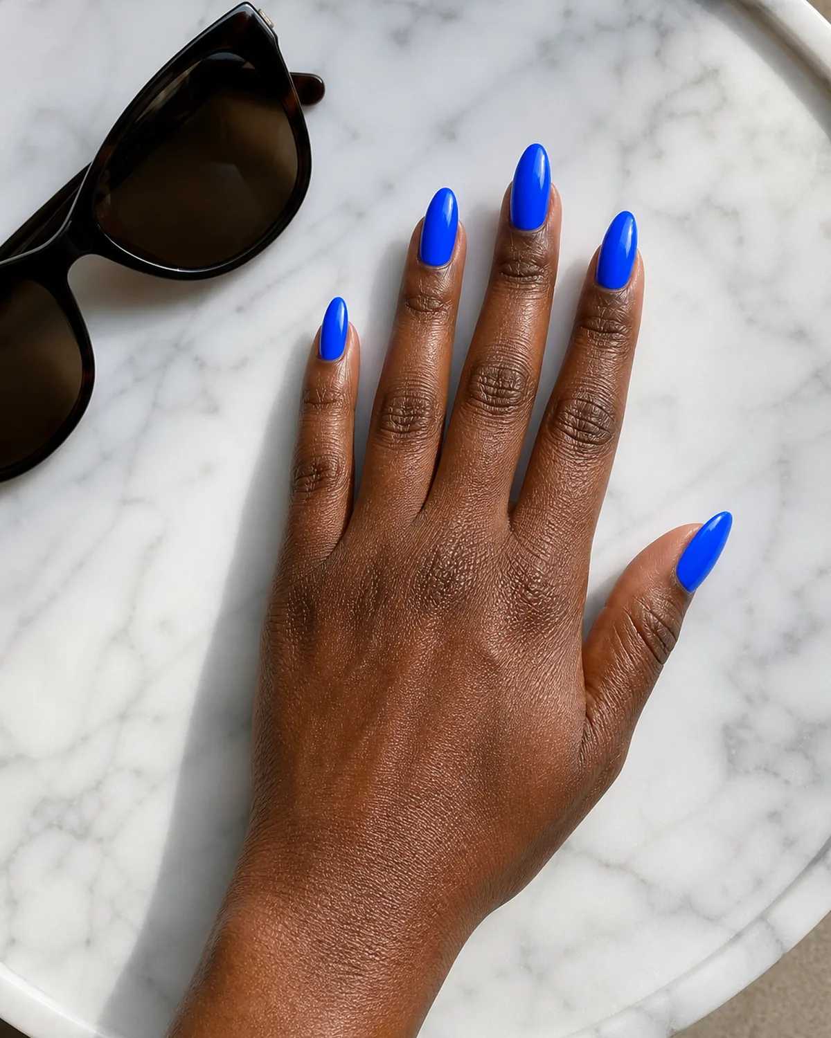 Single hand with bright neon electric blue nails on stiletto shape resting beside sunglasses on a white marble tray