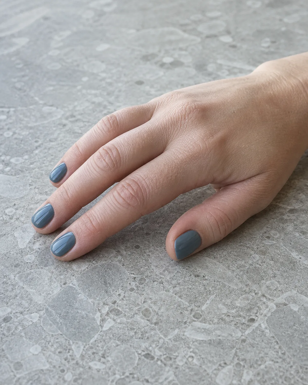 Single hand with steel blue gray-toned nails on short square shape lying relaxed on its side on a cool stone countertop
