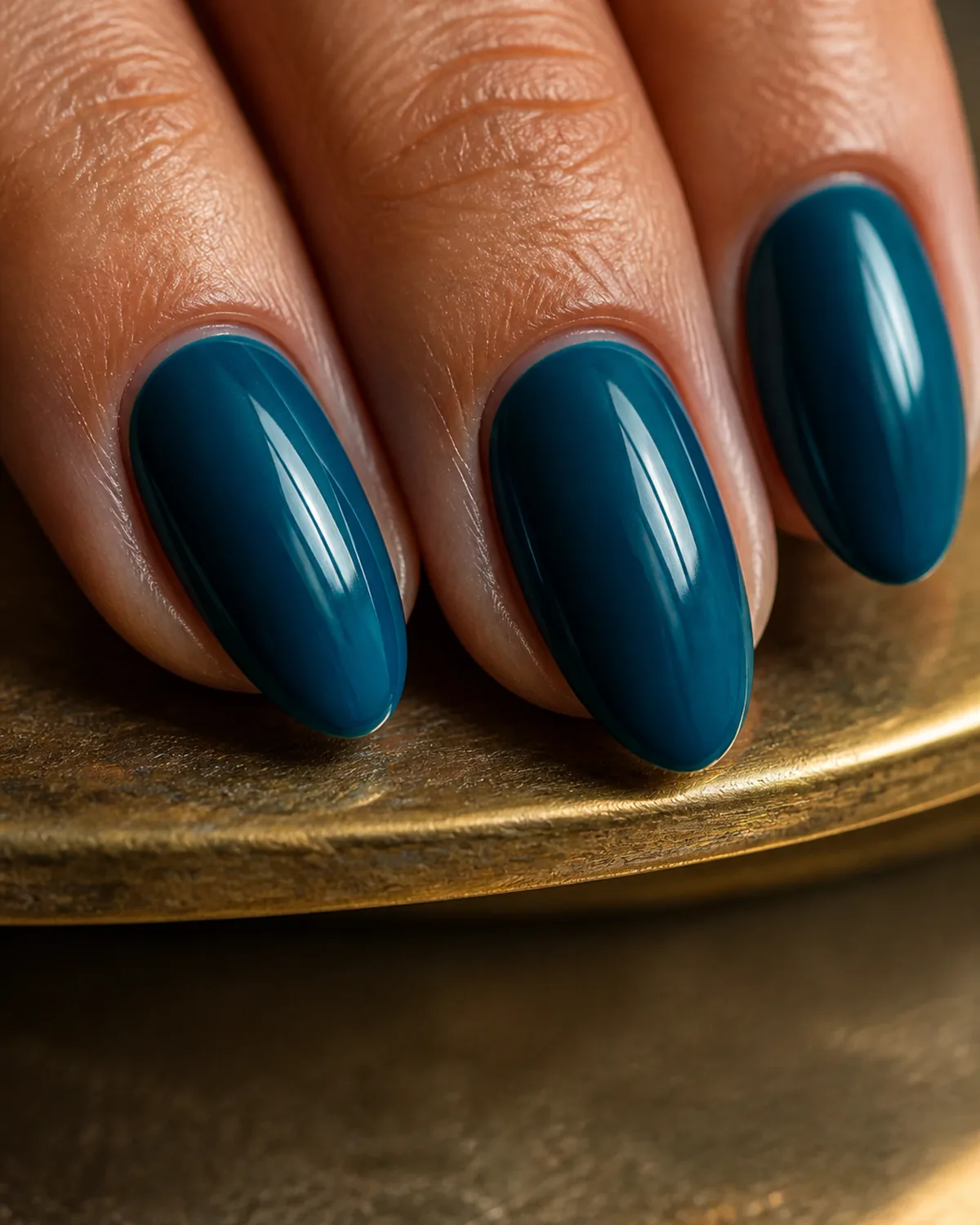 Extreme close-up macro of three fingertips with deep teal blue nails on almond shape resting on the edge of a brass tray
