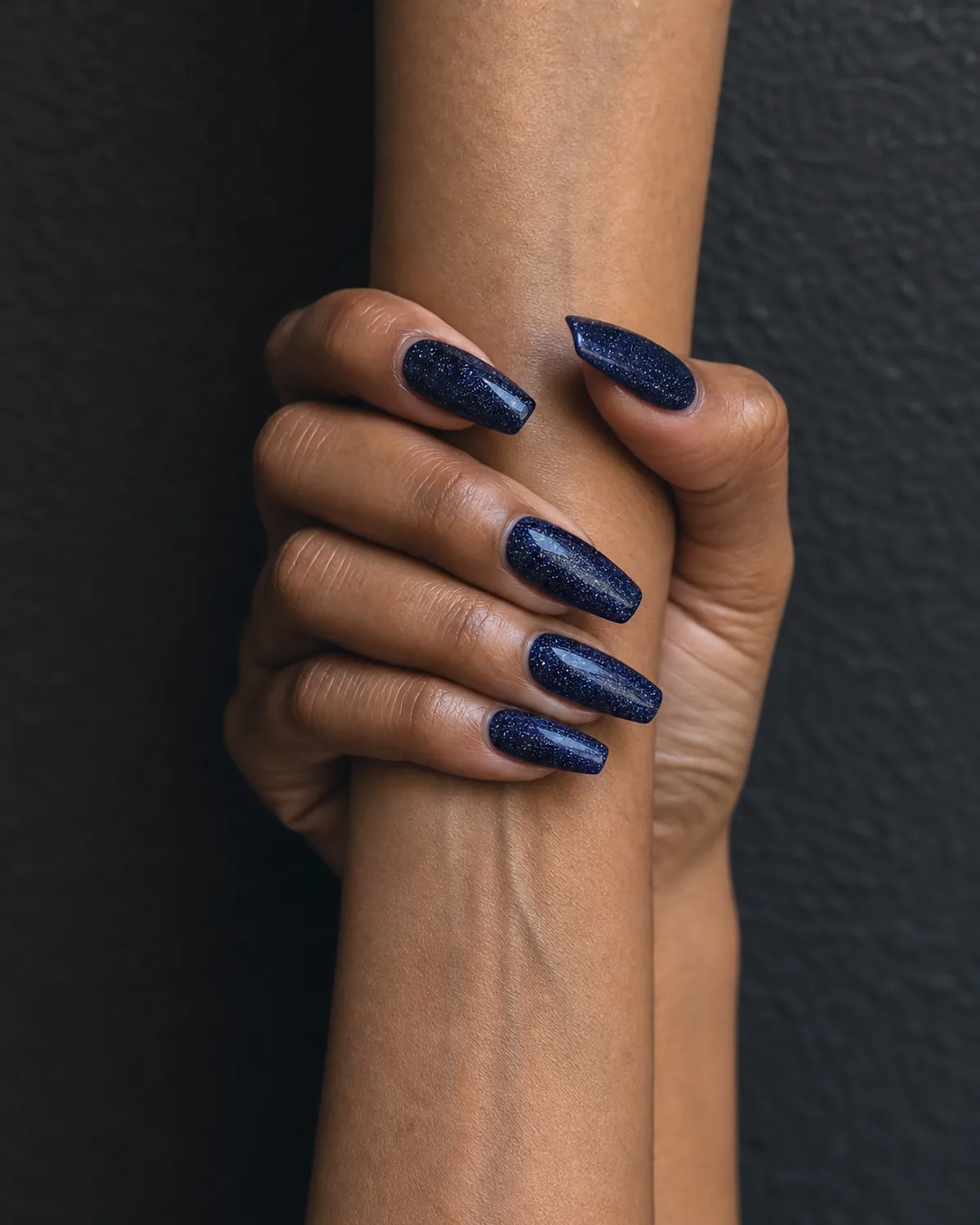 Single hand with midnight blue glitter nails on coffin shape loosely holding the opposite wrist against a dark wall