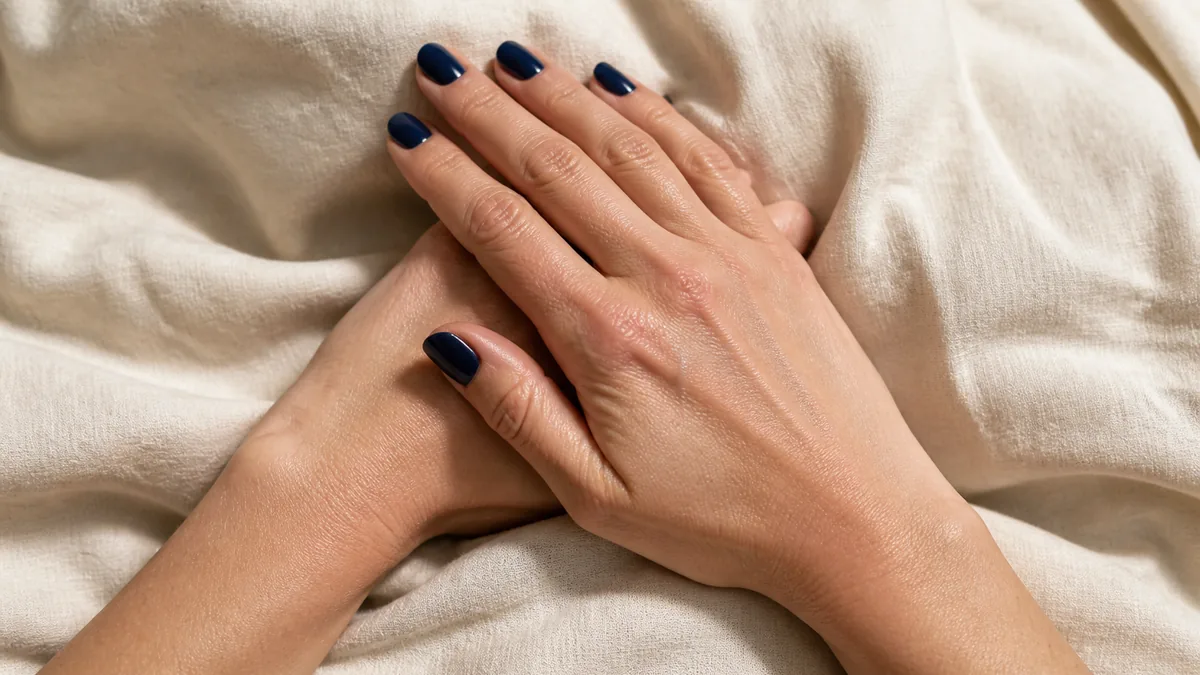 Two hands stacked with fingertips angled down showing deep navy blue nails on short almond shape resting on cream linen