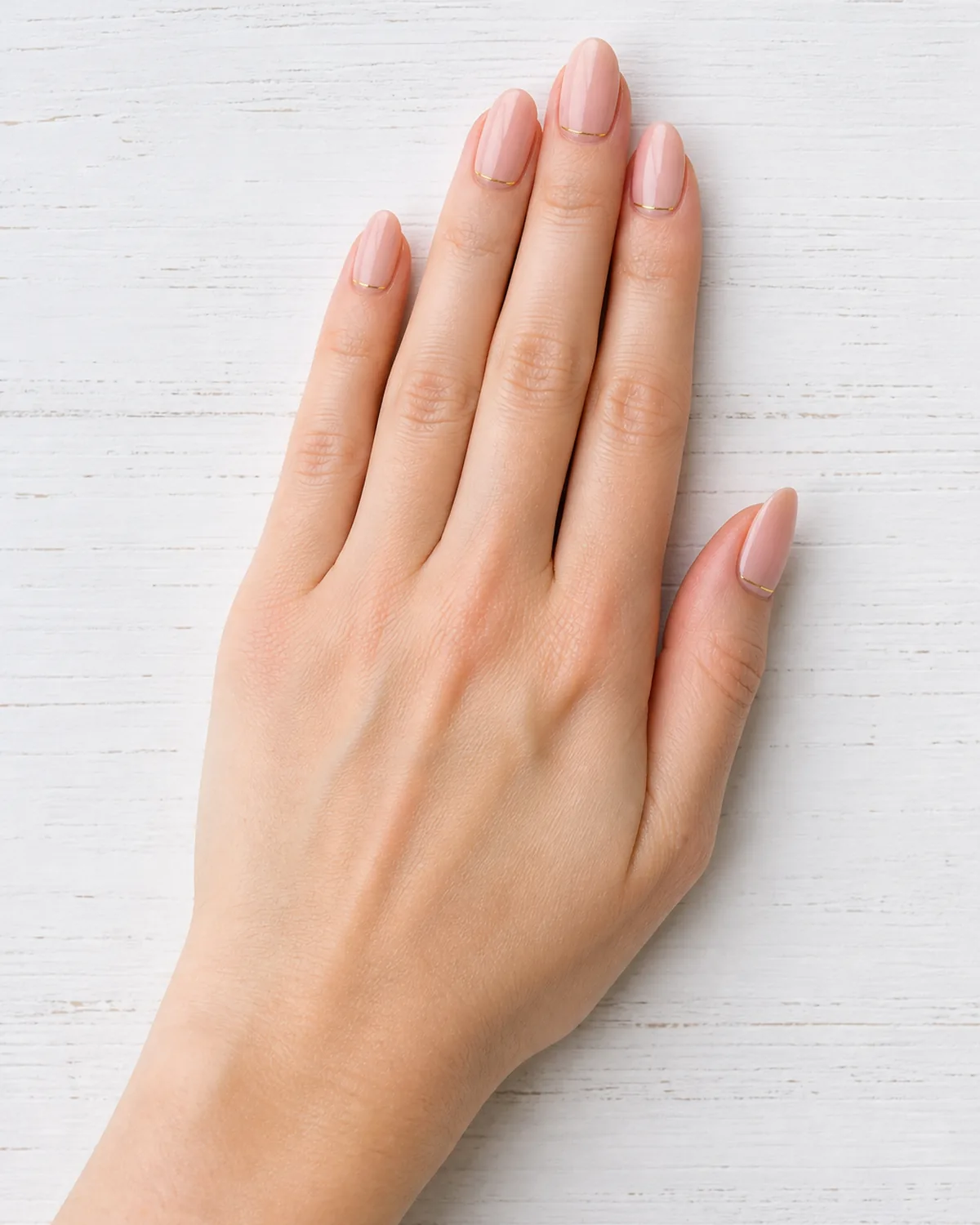 Single left hand with sheer pink nails and thin gold metallic cuff lines near the cuticle on medium almond shape palm down on white wood surface
