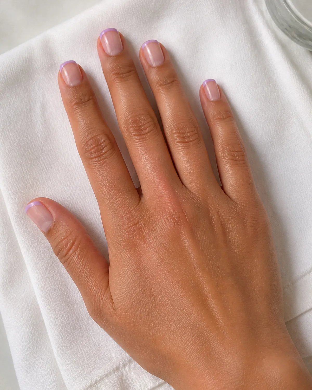 Single hand with sheer nude base and soft pastel lilac French tips on short square nails on folded white cotton napkin near clear glass edge