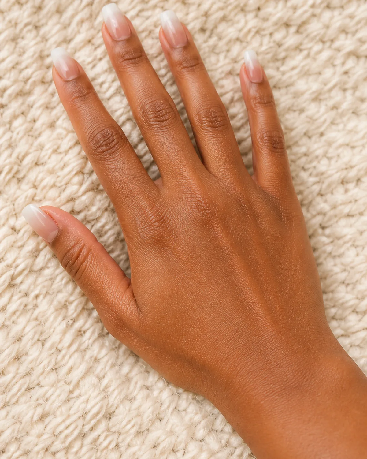Single hand with warm nude to white ombre gradient on medium coffin nails palm down on cream knit texture