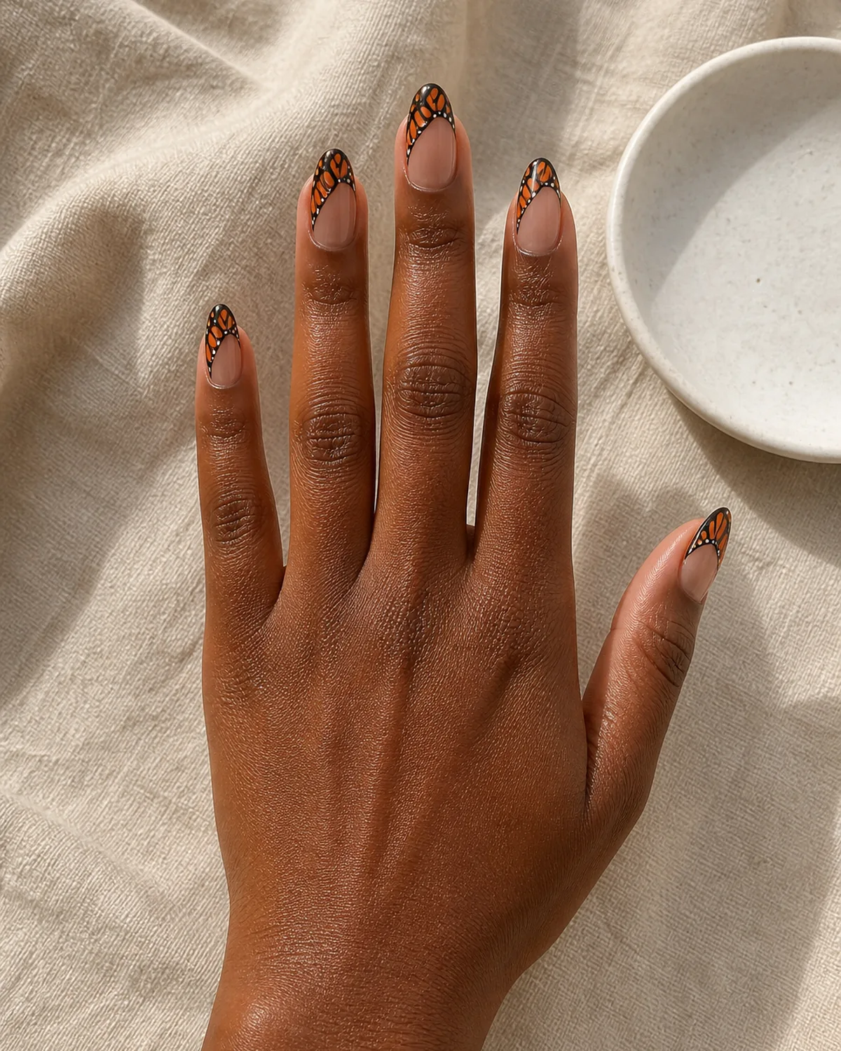 Single hand with sheer nude nail polish on almond nails with hand-painted orange and black monarch butterfly wing patterns extending from the tip inward resting palm-down on warm beige linen surface