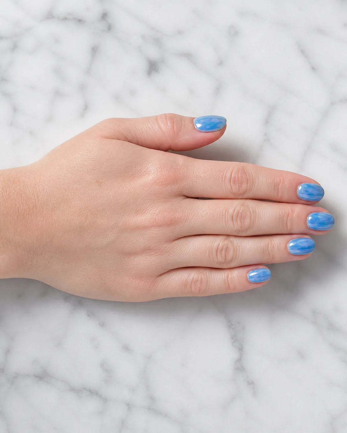 Single hand with translucent iridescent blue watercolor wash nail polish on short oval nails mimicking blue morpho butterfly wing shimmer resting flat on white marble countertop