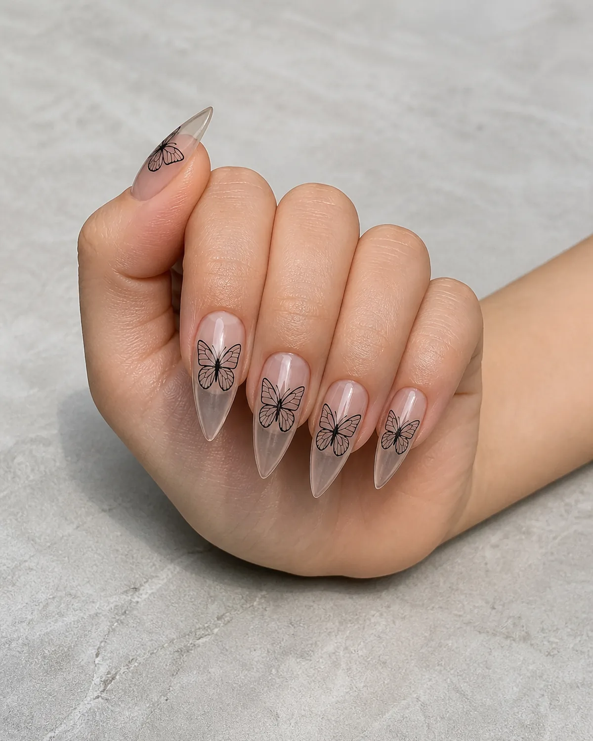 Single hand with clear bare stiletto nails with single black outline butterfly drawn on each nail with wings open and no fill resting on pale gray stone surface