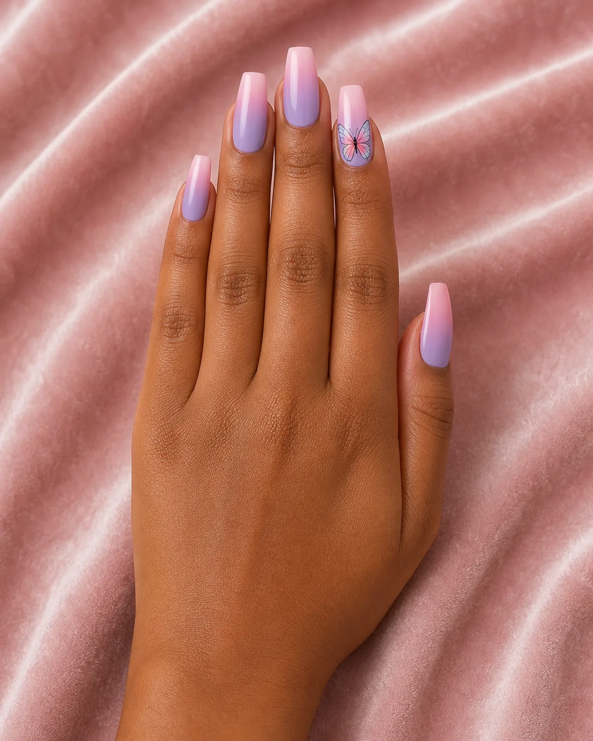 Single hand with soft lavender-to-pink ombre nail polish on coffin nails with one accent nail featuring detailed pastel butterfly resting on blush-pink velvet surface