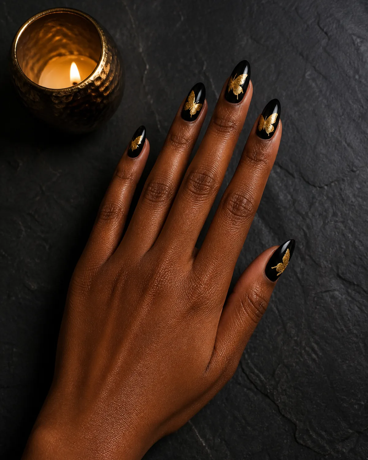 Single hand with glossy black nail polish on almond nails with gold metallic foil butterfly silhouettes pressed onto each nail resting on dark charcoal slate surface beside small gold tealight holder