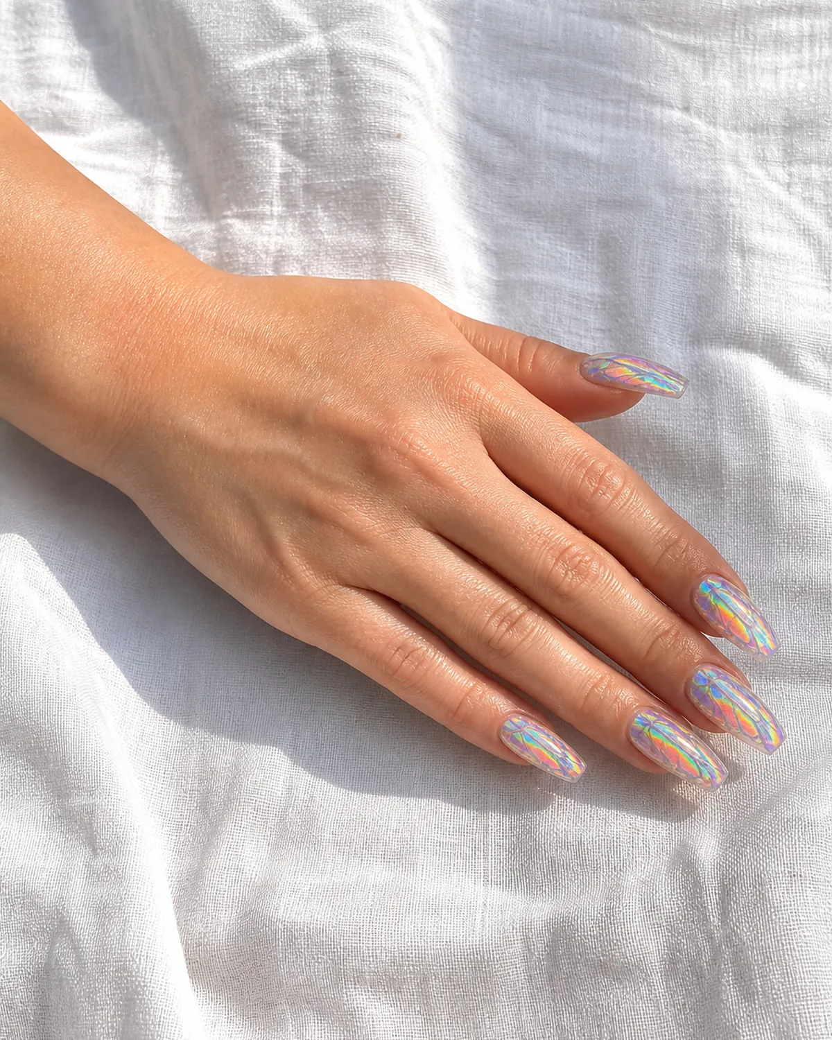 Single hand with clear holographic duochrome top coat nail polish on long coffin nails with butterfly wing shapes visible in light shift resting on white textured linen surface