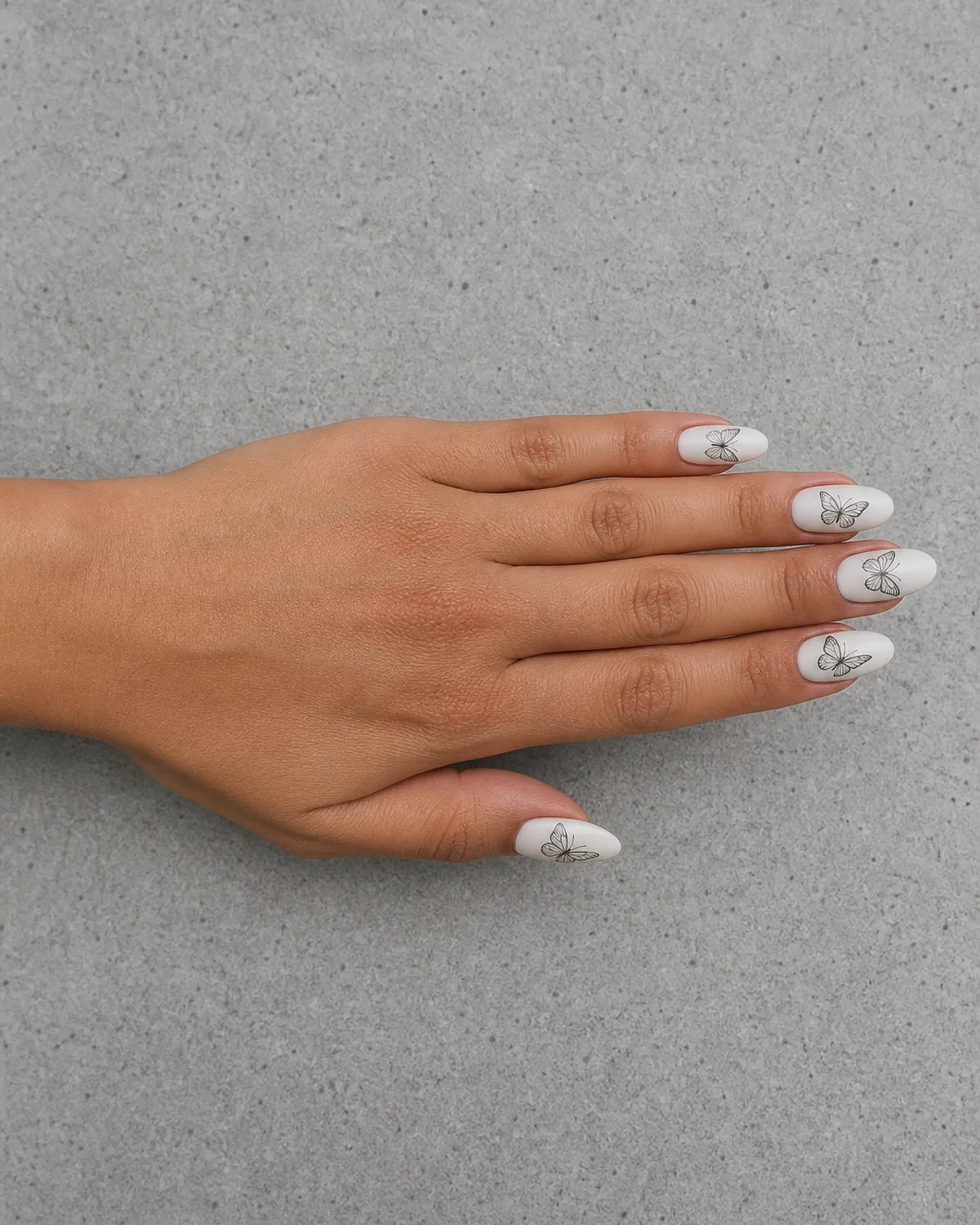 Single hand with matte white nail polish on almond nails with fine black line-drawn butterflies in different flight poses across each nail resting on light gray concrete surface