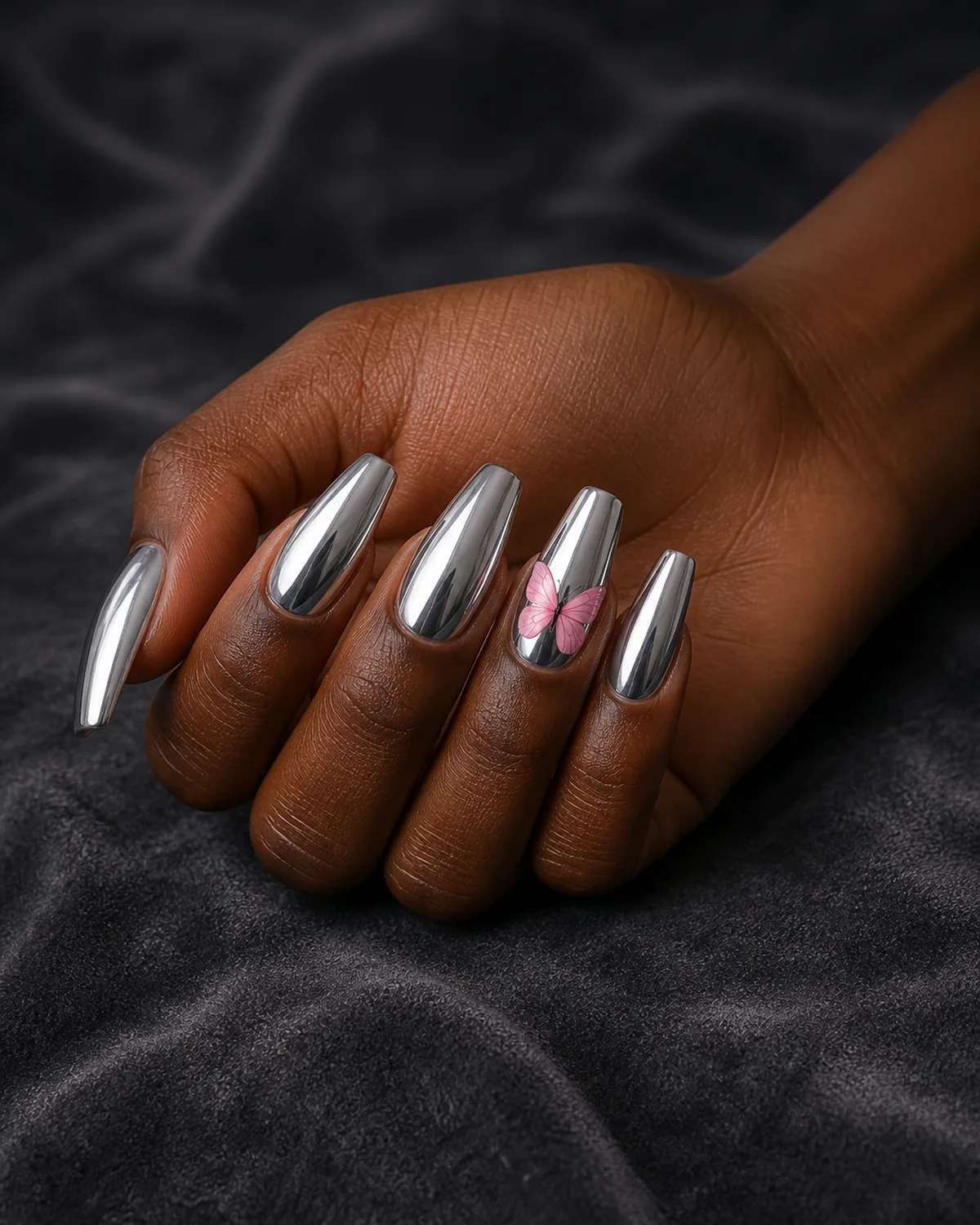Single hand with chrome silver mirror finish nail polish on coffin nails with soft pink butterfly painted on one accent nail resting on dark gray velvet surface