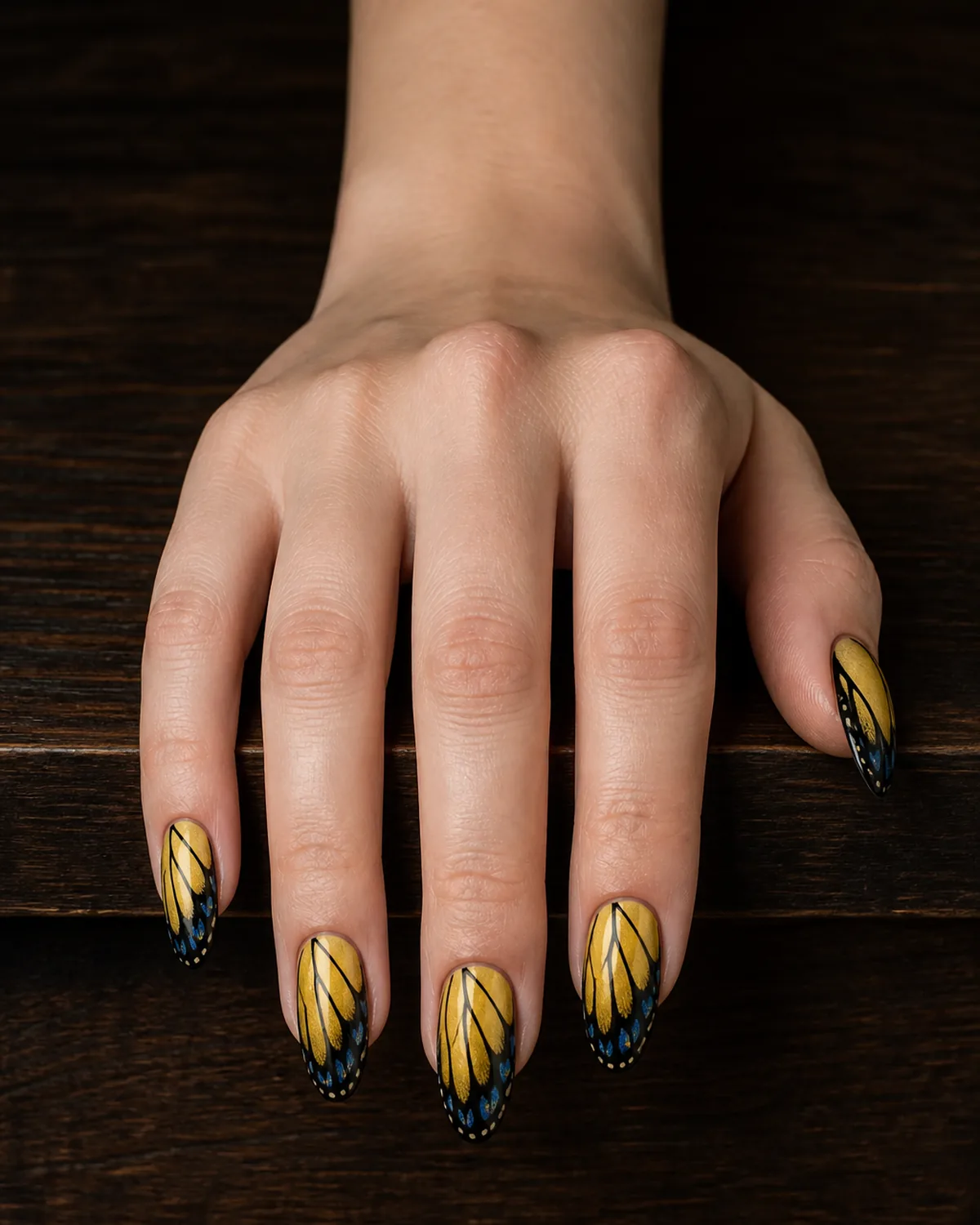 Single hand with yellow fading to black gradient nail polish on long oval nails with blue accent spots mimicking eastern tiger swallowtail butterfly wing pattern resting on dark wood table