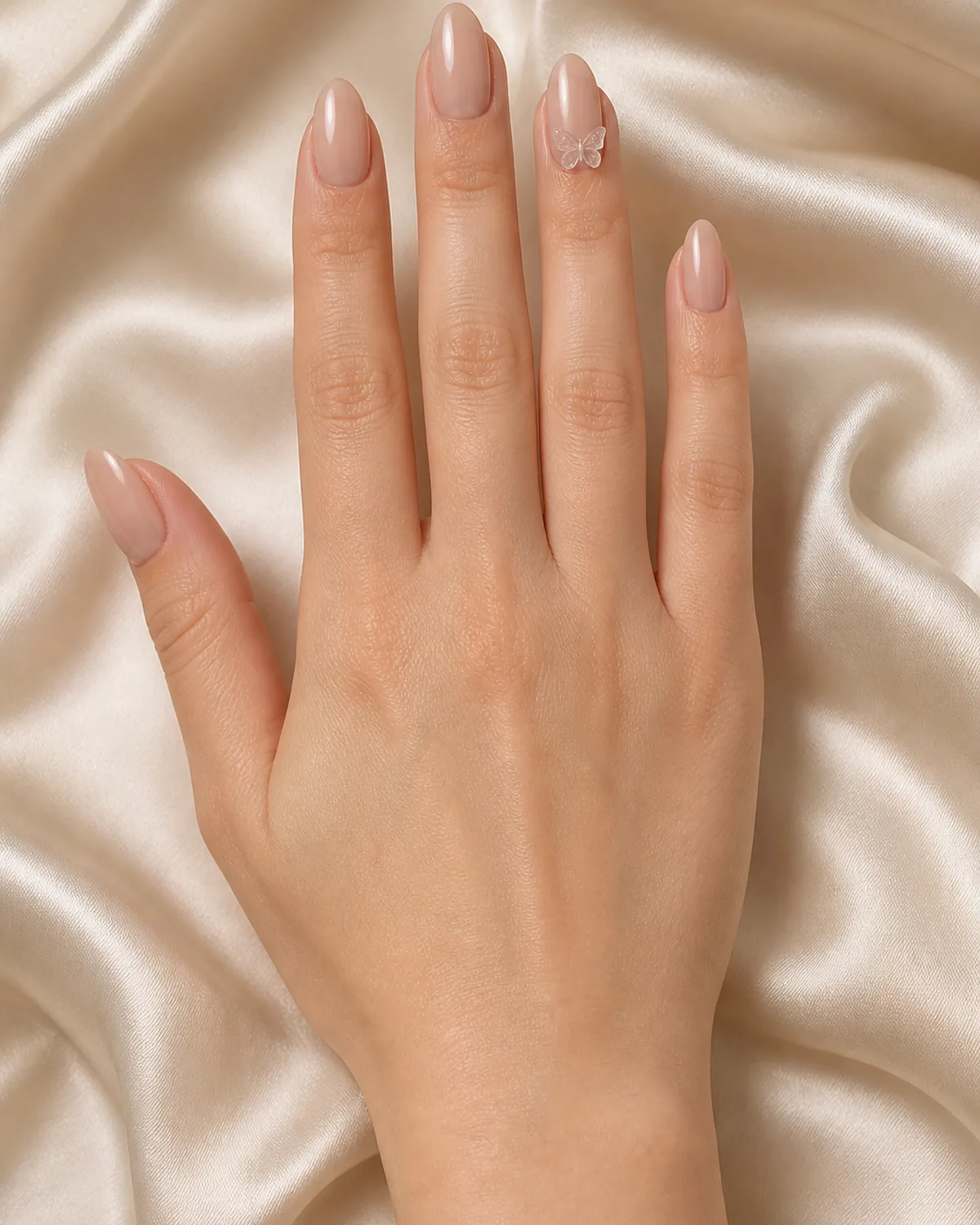 Single hand with sheer nude nail polish on almond nails with small 3D resin butterfly charm adhered near cuticle on ring finger resting palm-down on cream satin fabric surface