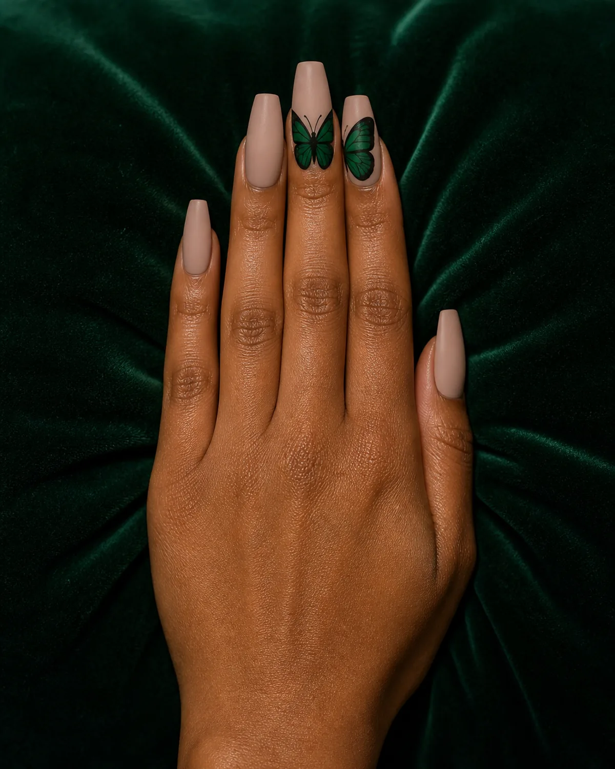 Single hand with matte nude nail polish on coffin nails with rich emerald green butterfly painted across two adjacent nails ring and middle finger forming continuous wing span resting on dark green velvet cushion