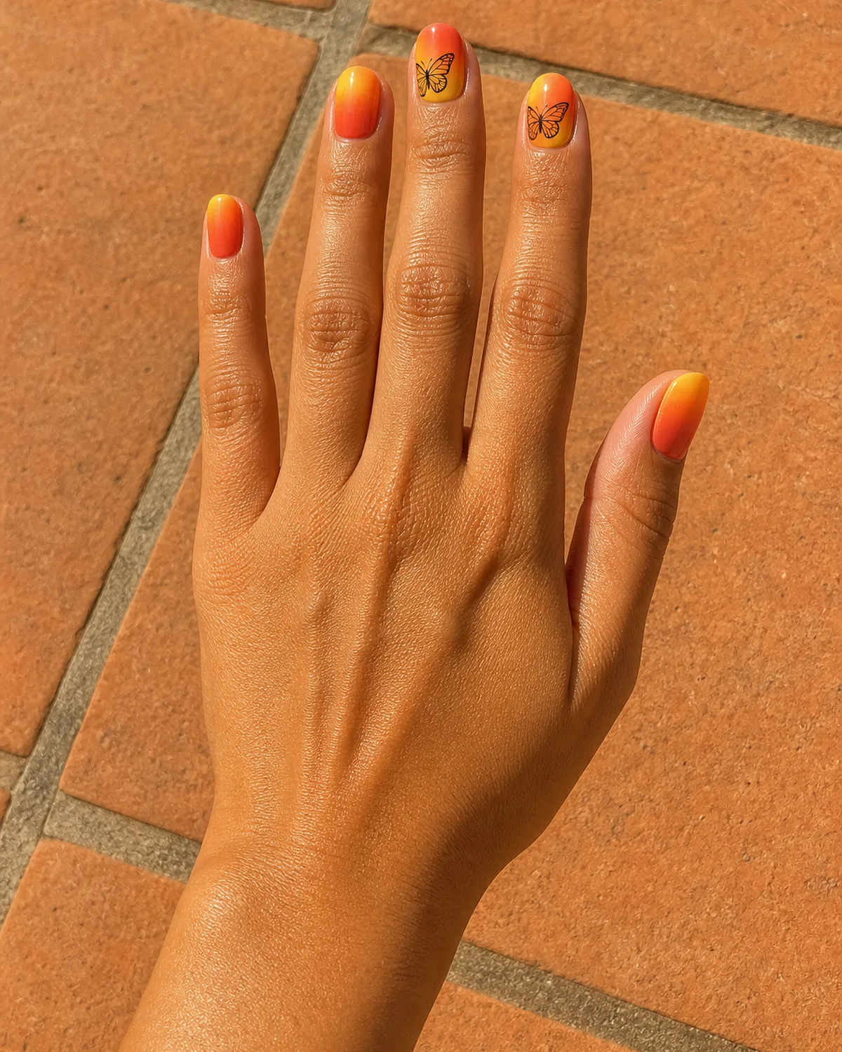 Single hand with warm sunset gradient nail polish golden yellow to coral to deep orange on round nails with delicate black butterfly outlines on two accent nails resting on warm terracotta tile surface