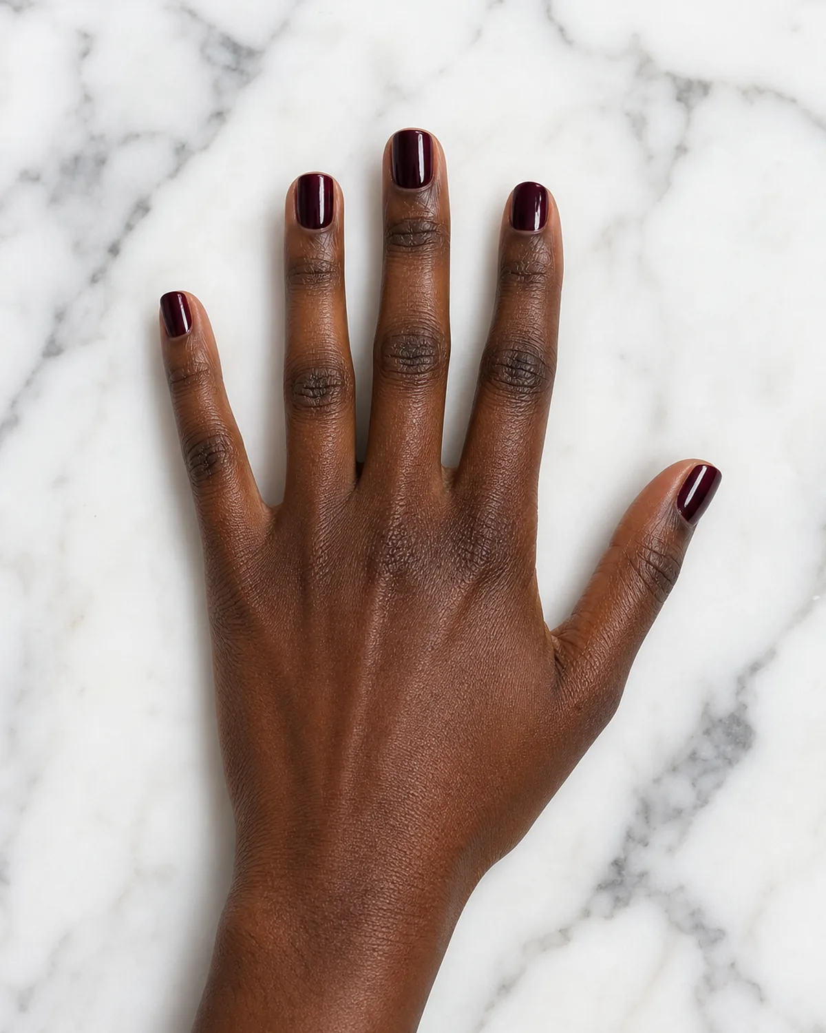 Single hand splayed flat on white marble showing deep burgundy polish on short square nails